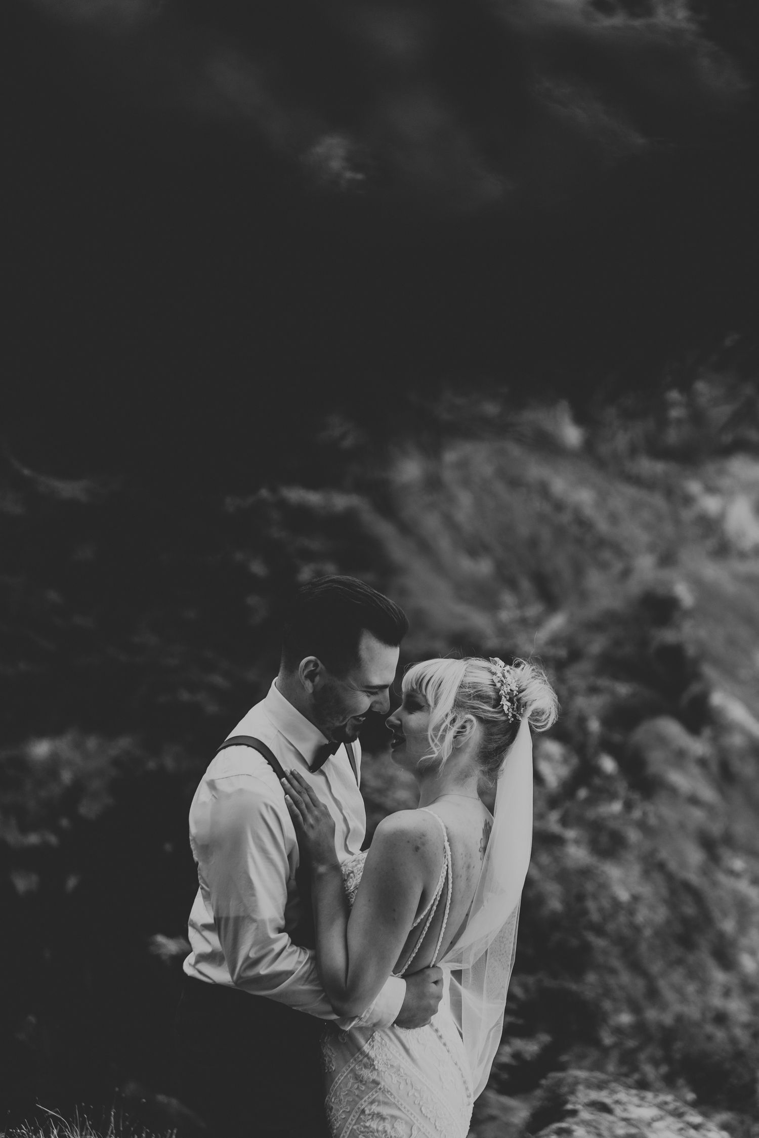 A dramatic black and white wedding portrait shows a romantic moment between a couple against a mountainous backdrop.