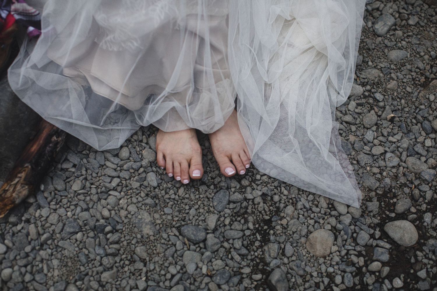 Close-up of barefoot bride standing on gravel in a flowing tulle wedding gown.