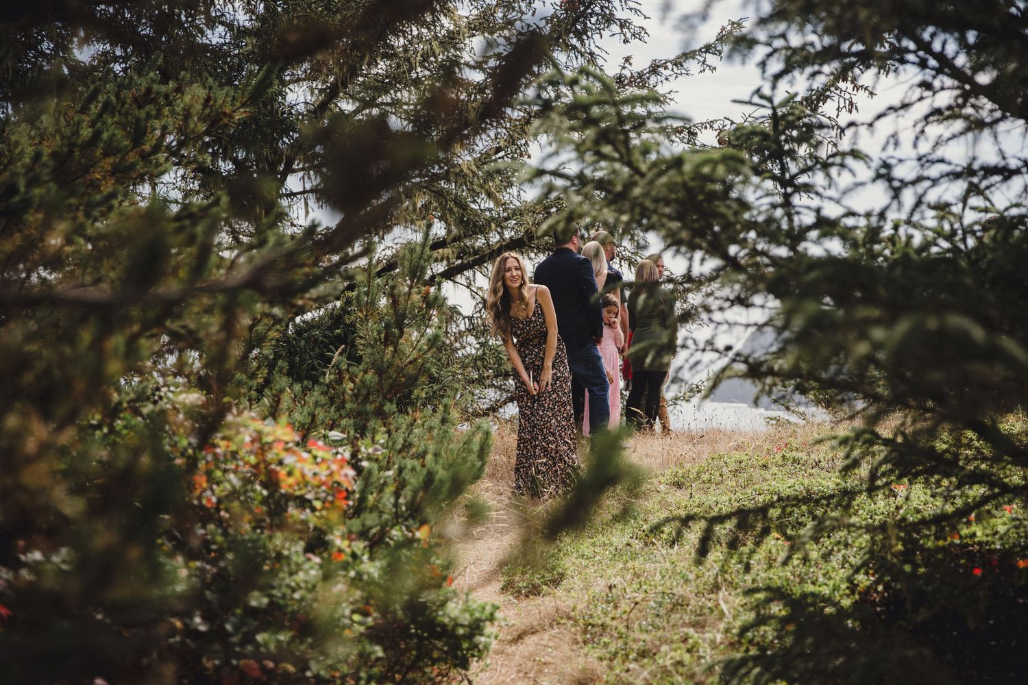 Three people walking along a scenic garden path framed by lush green trees and coastal wildflowers.