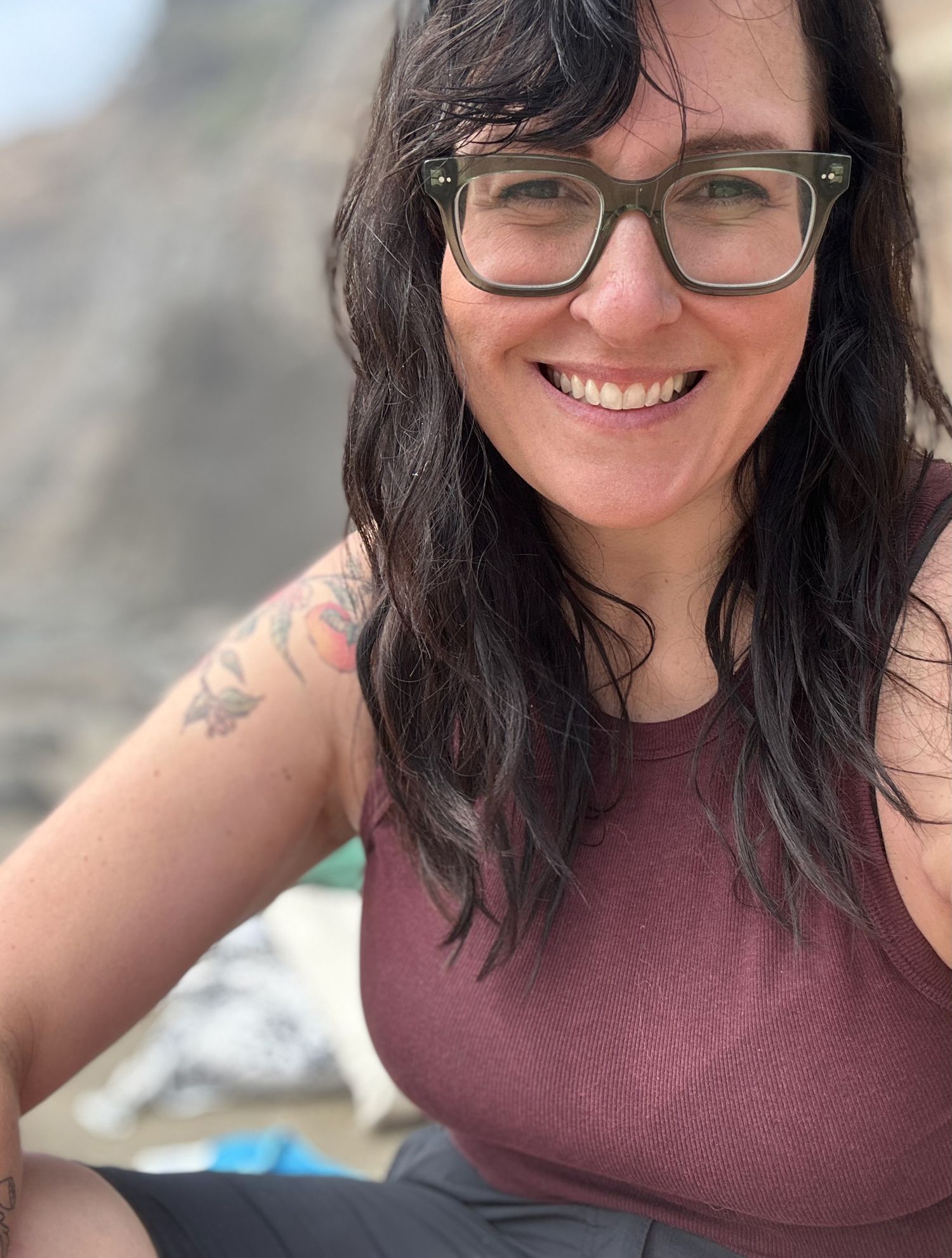 Smiling woman with dark hair, green glasses, and floral shoulder tattoo wearing a maroon tank top outdoors.