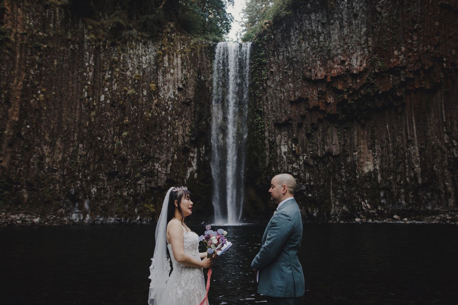 Couple exchanges vows in front of a dramatic waterfall surrounded by rocky cliffs.