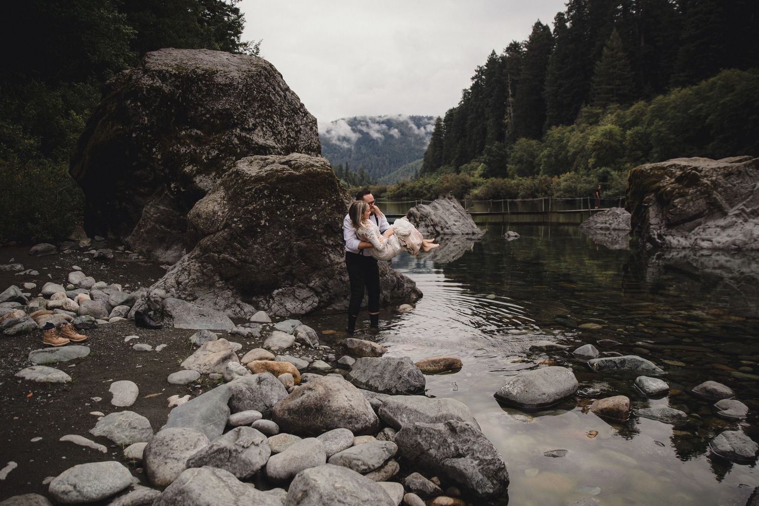 Couple embracing on rocky riverbank surrounded by lush forest and misty mountains in a scenic wilderness elopement.