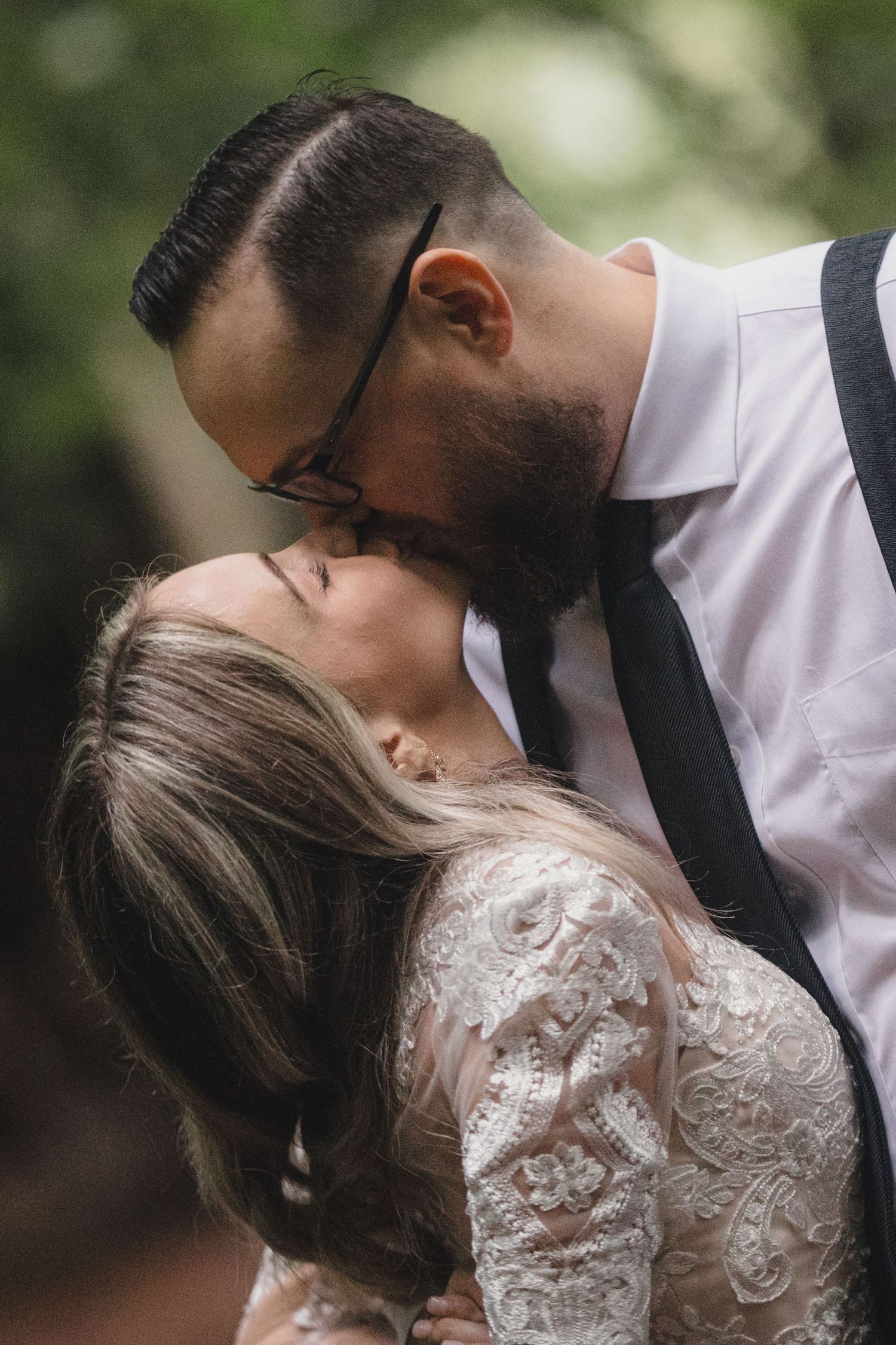 Bearded groom with glasses kisses bride in lace wedding dress in a romantic outdoor portrait.