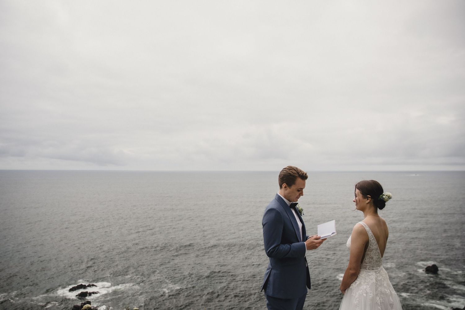 Bride and groom exchange vows on a dramatic coastal cliff overlooking a grey ocean on their wedding day.