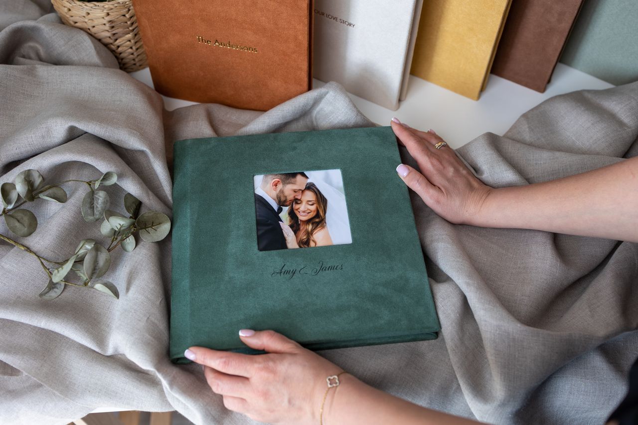 Hands holding a green velvet wedding photo album with a couple's portrait on the cover, surrounded by fabric and greenery.