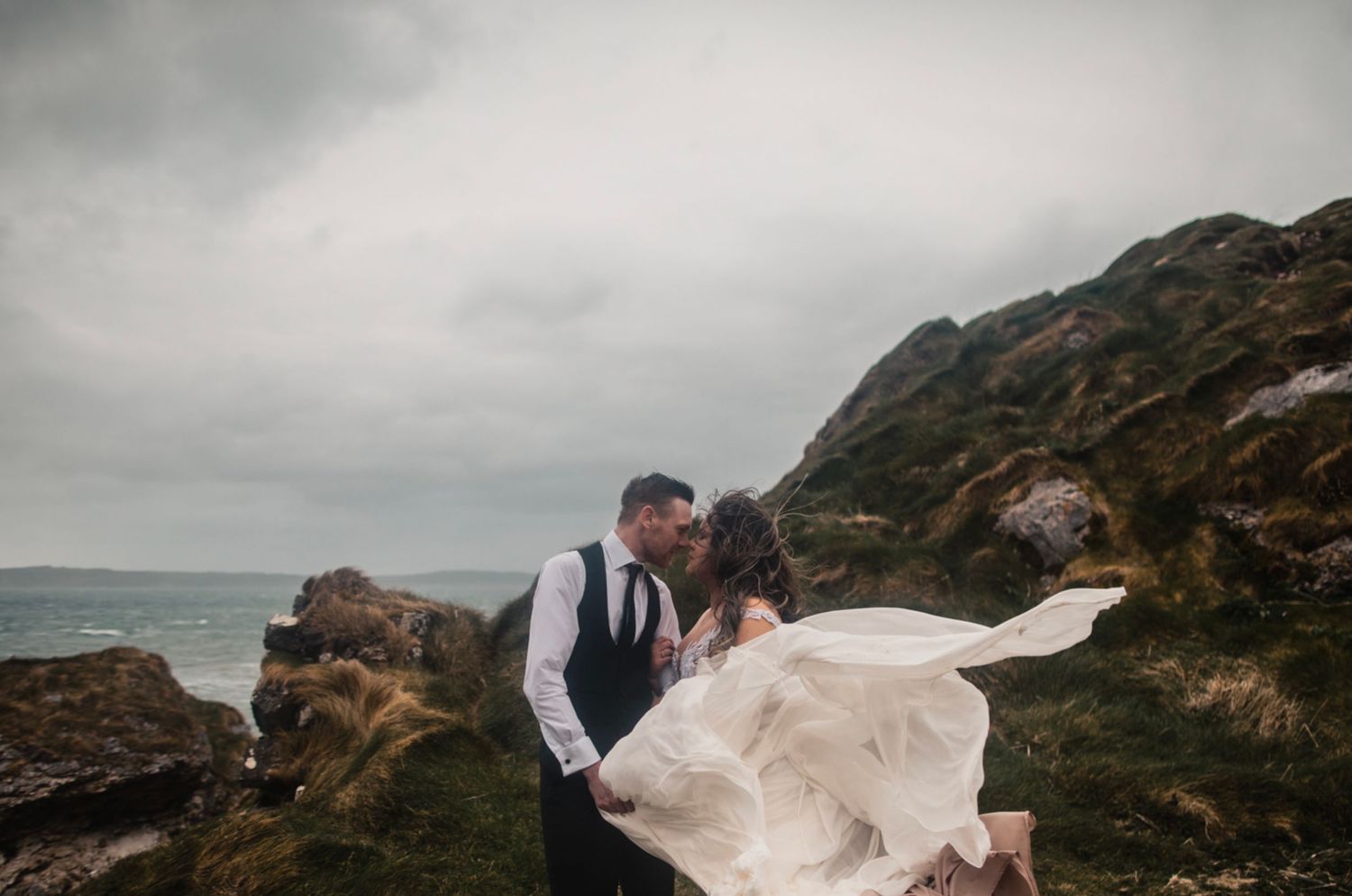 A wedding couple embraces on dramatic coastal cliffs with windswept dress and moody stormy skies.