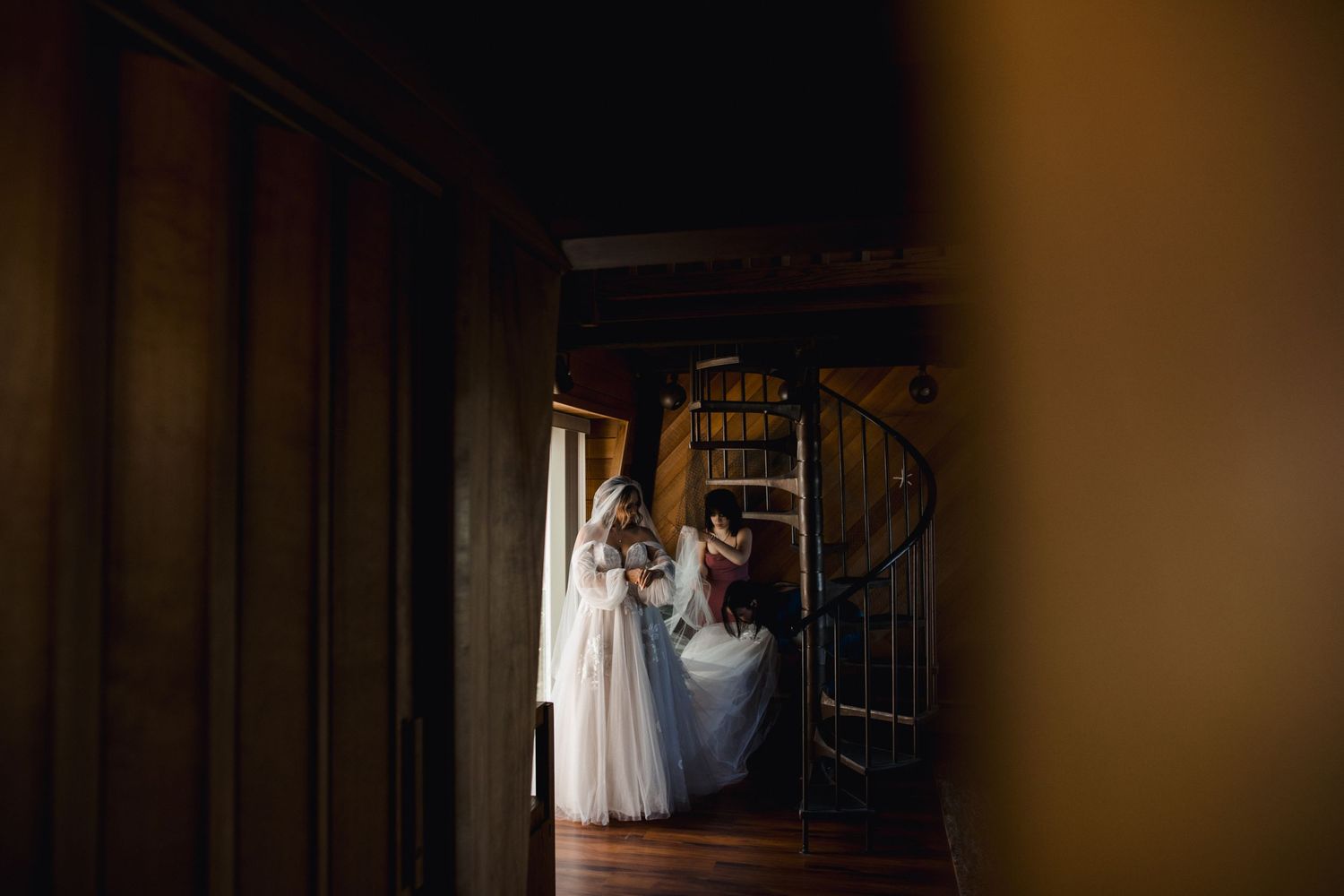 Wedding dress hangs in doorway with spiral staircase visible in soft, moody lighting.