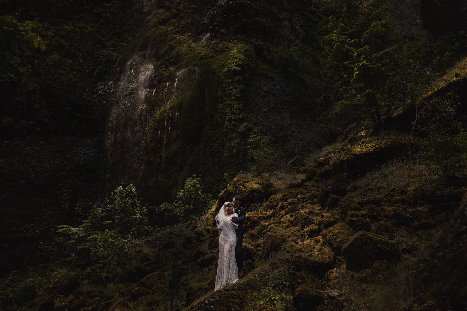 A bride in white stands dramatically against a dark forest cliff backdrop.
