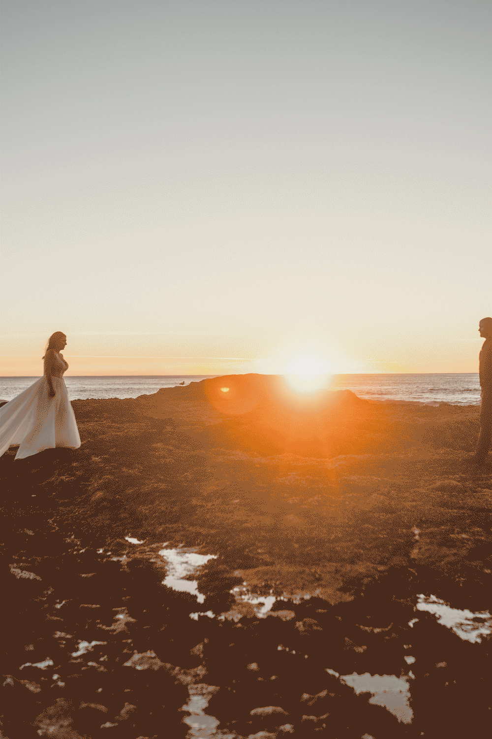 A dramatic coastal sunset silhouettes a figure walking along a rocky beach with waves crashing against the shore.