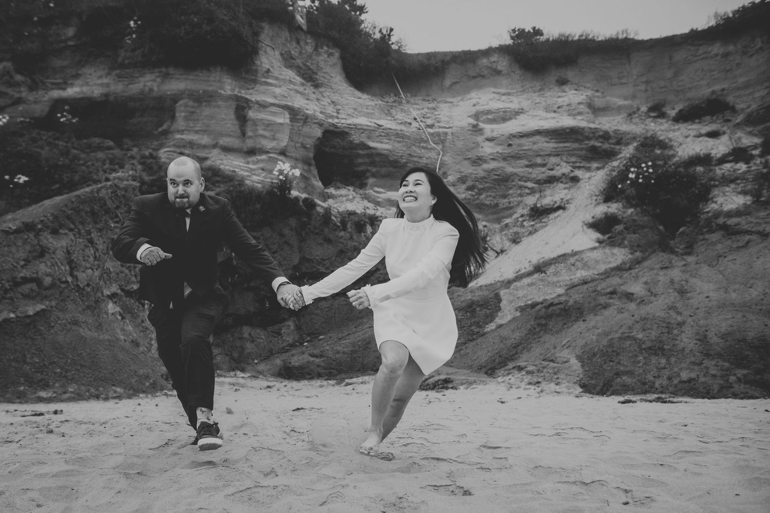 Black and white photo of couple running hand in hand on sandy beach.