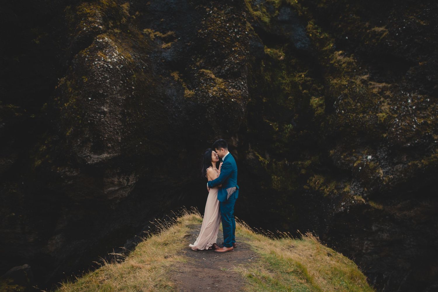 Couple shares intimate moment on grassy hilltop with dramatic mountains in background.