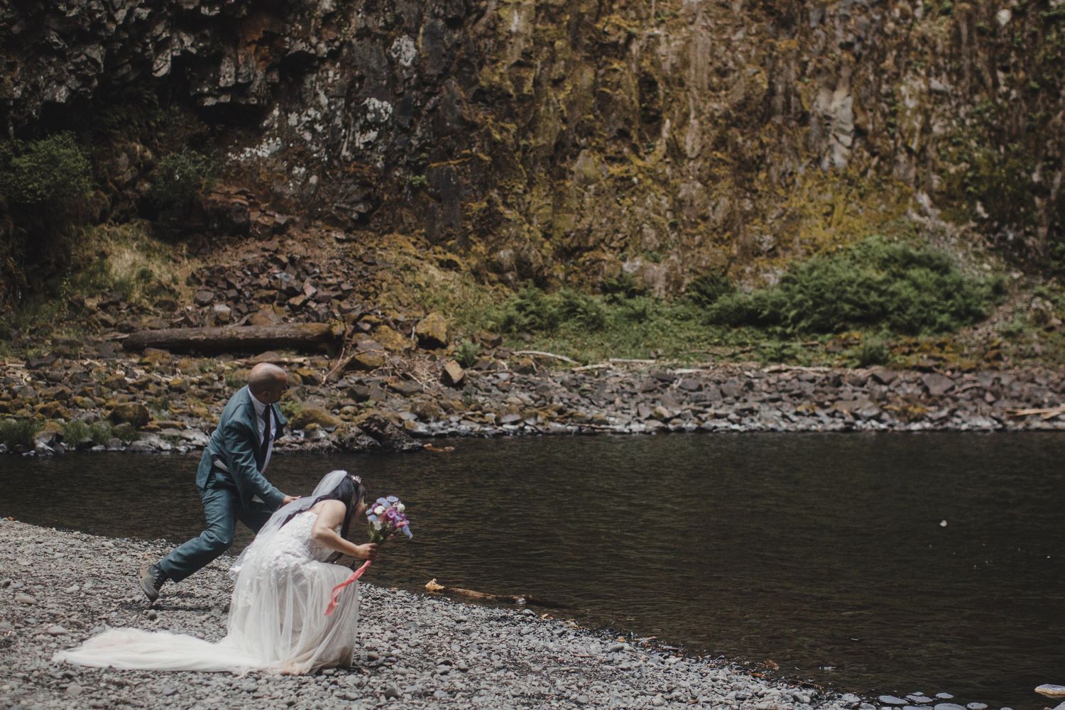 A bride and groom share a romantic moment on a rocky riverbank beneath towering cliffs.