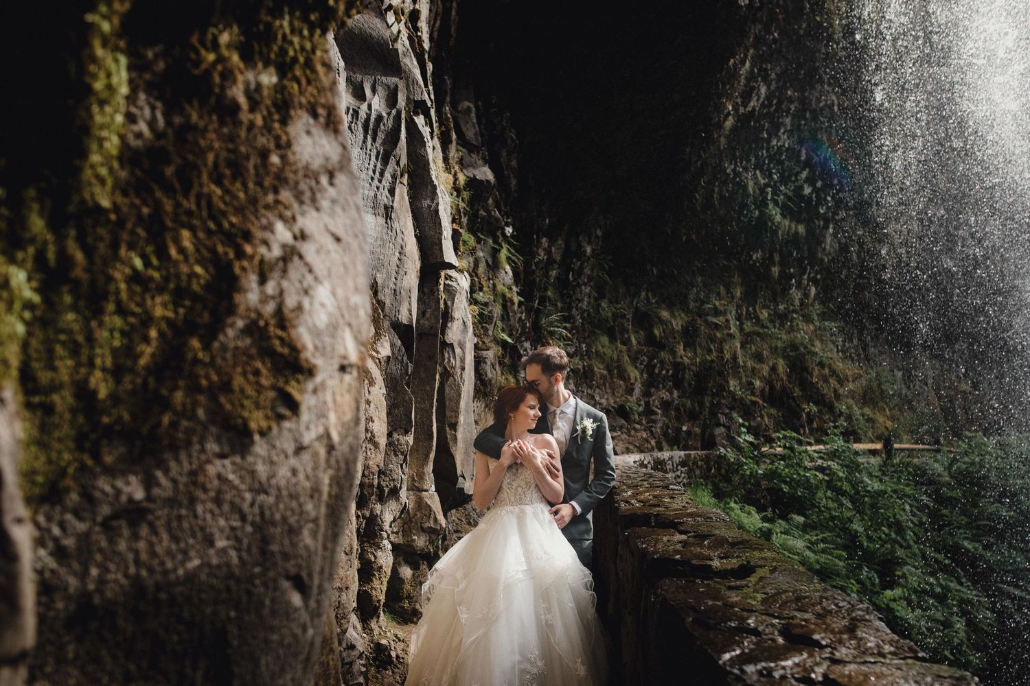 Couple in formal wedding attire pose in a dramatic cave setting with natural light streaming through moss-covered walls.
