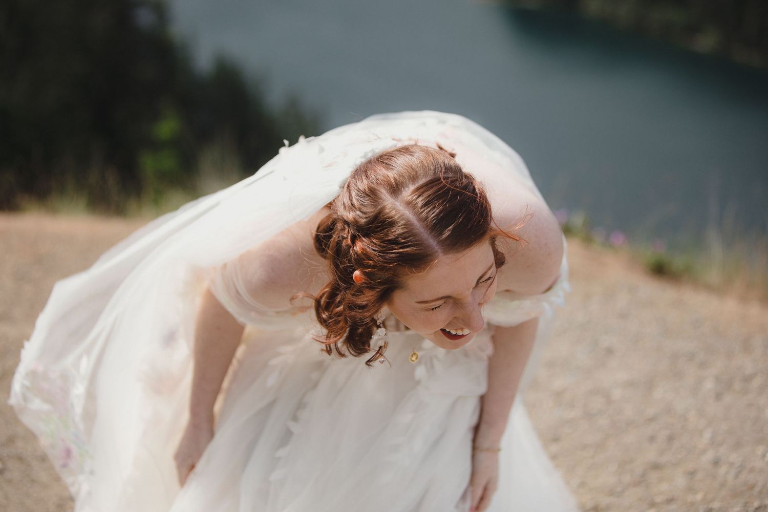 Bride in white dress with flowing veil bends down laughing at Diable lake in washington.