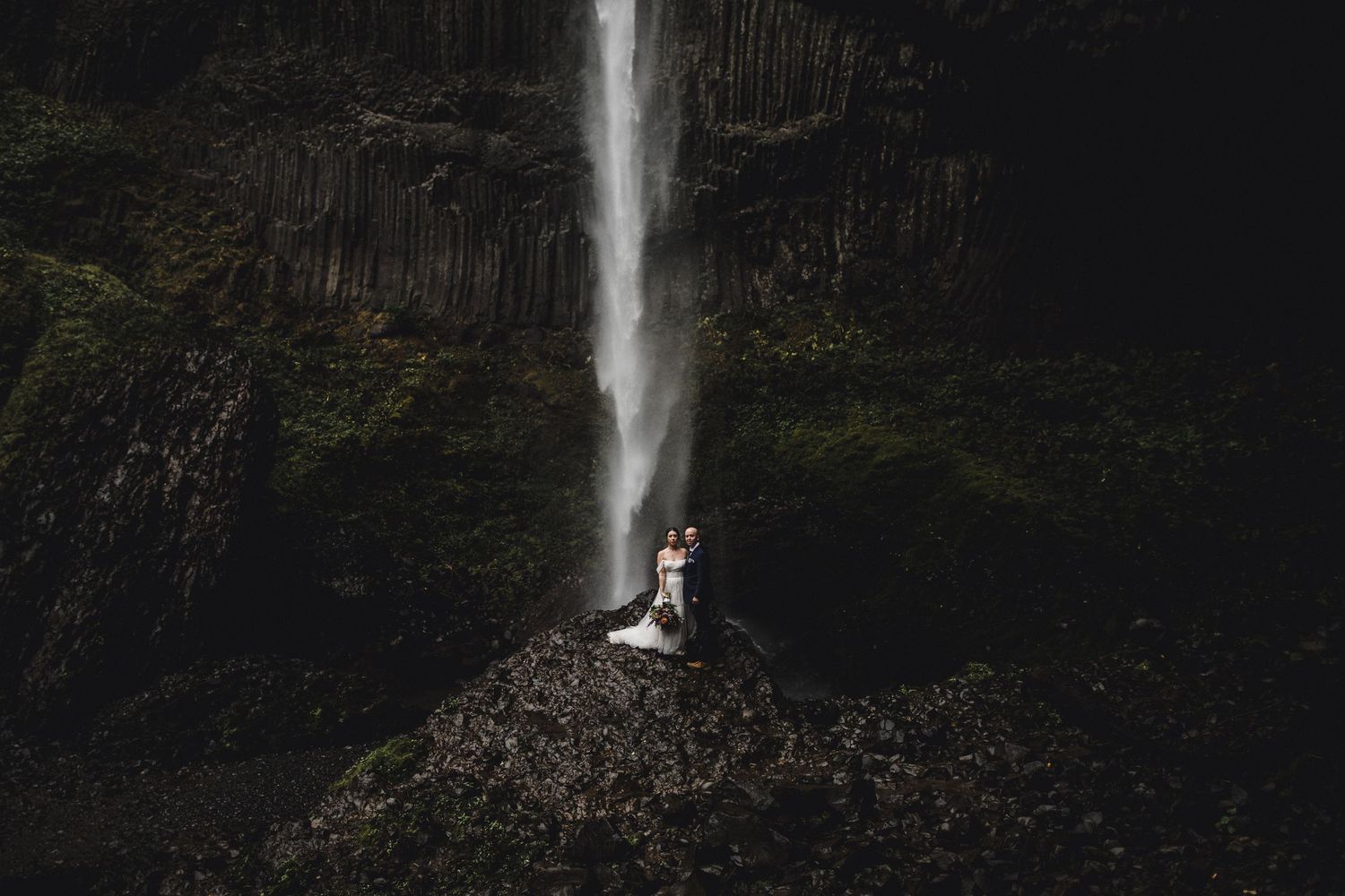Couple embracing on rocky ledge before dramatic waterfall with dark basalt columns in moody elopement photo.
