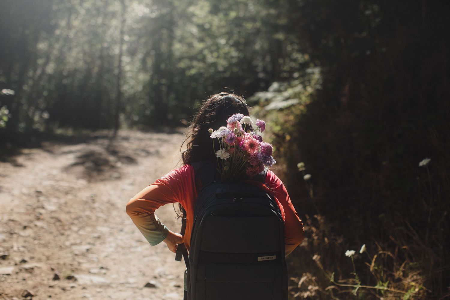 Woman with floral hair accessory and red jacket hiking on a sunlit forest trail.