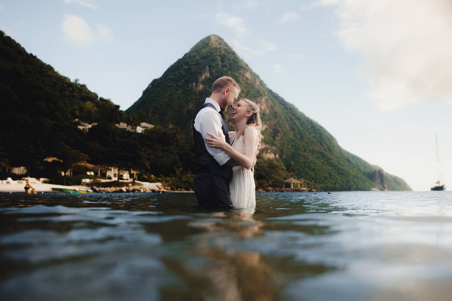 Couple sharing a romantic kiss while standing in shallow ocean water with lush green mountains in the background.