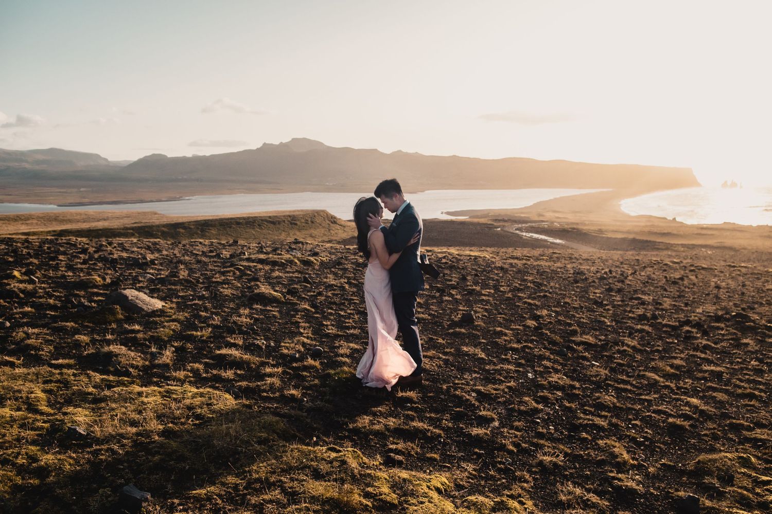 Two silhouettes share an intimate moment against a dramatic coastal sunset landscape.