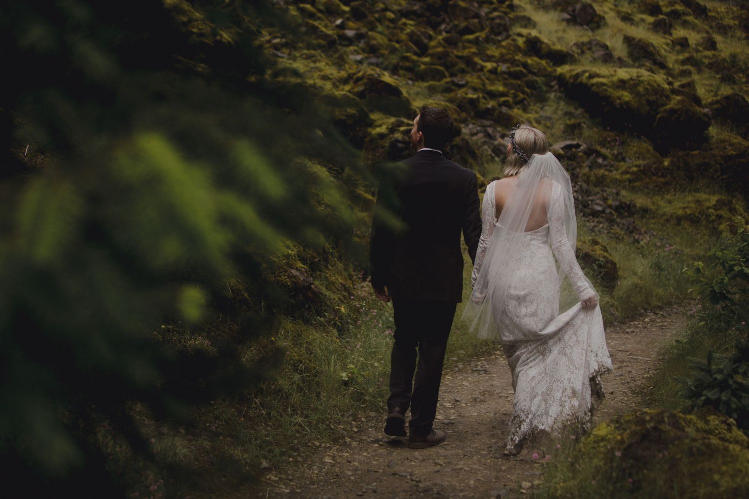 Newlyweds walking along a mossy mountain path with the bride's dress trailing behind.