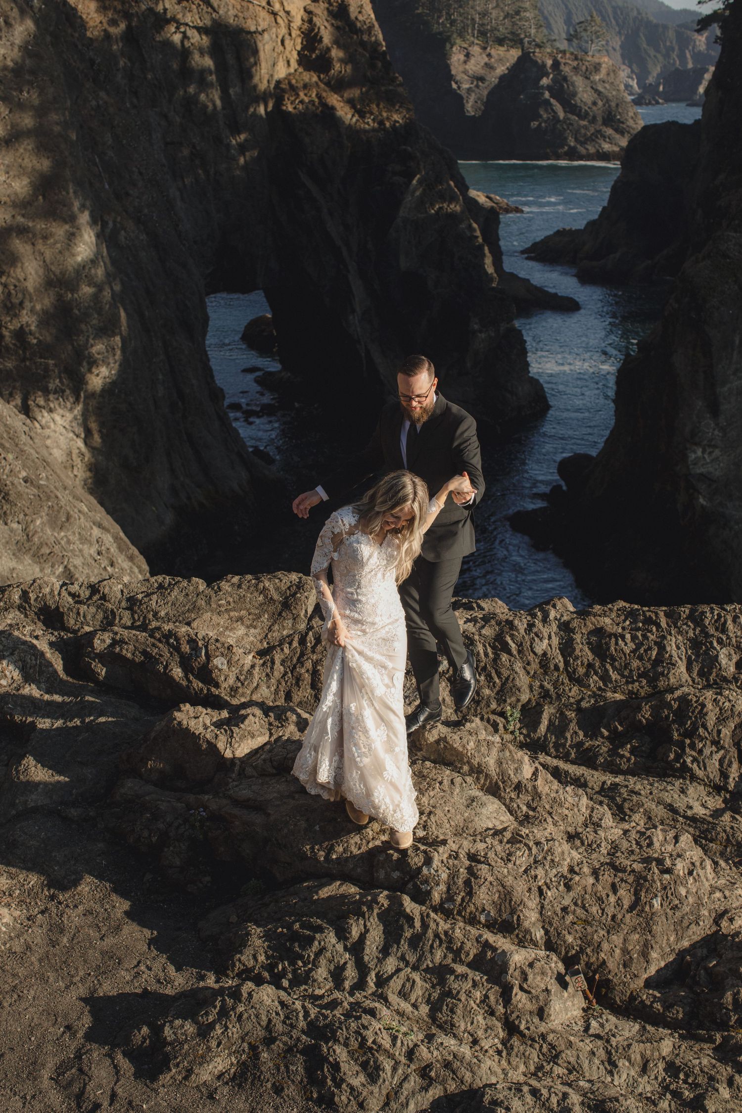 Bride and groom carefully climb rugged coastal rocks with dramatic sea stacks and ocean waves below.
