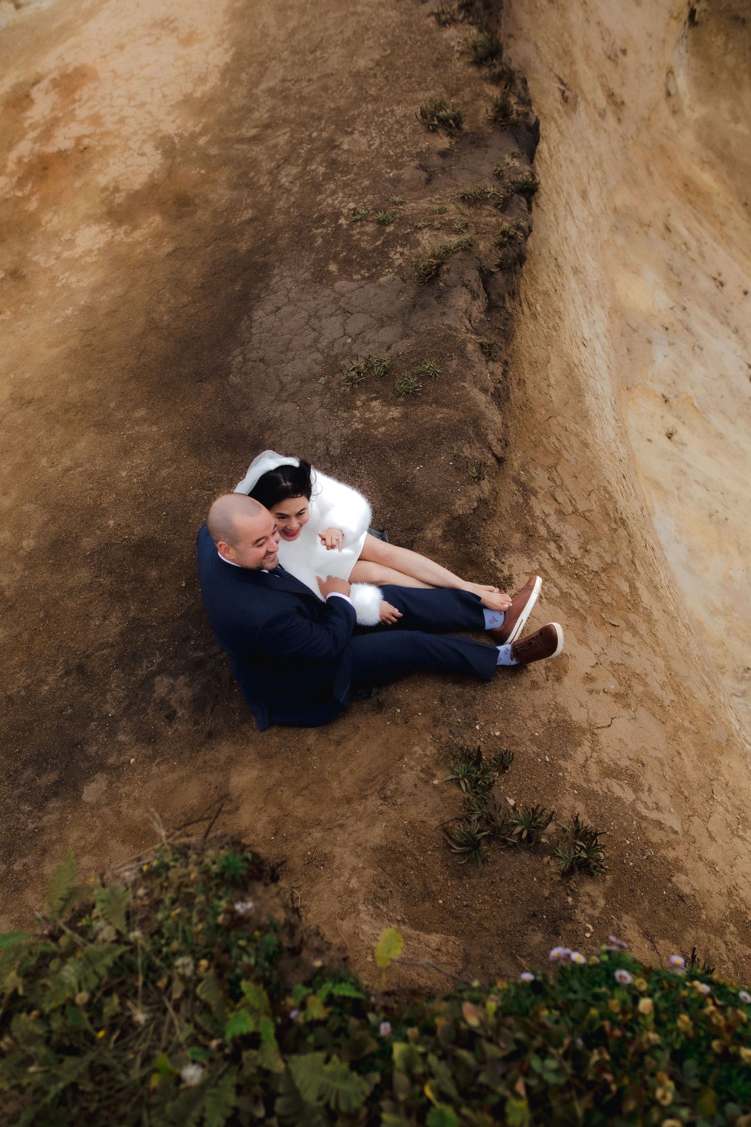 Aerial view of bride and groom sitting together on sandy ground during outdoor wedding.