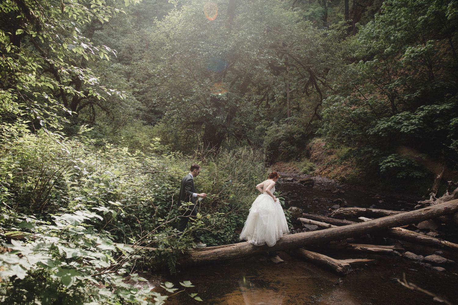 Bride and groom in wedding attire crossing a rustic log bridge over a stream in a lush green forest at Silver Falls State Park
