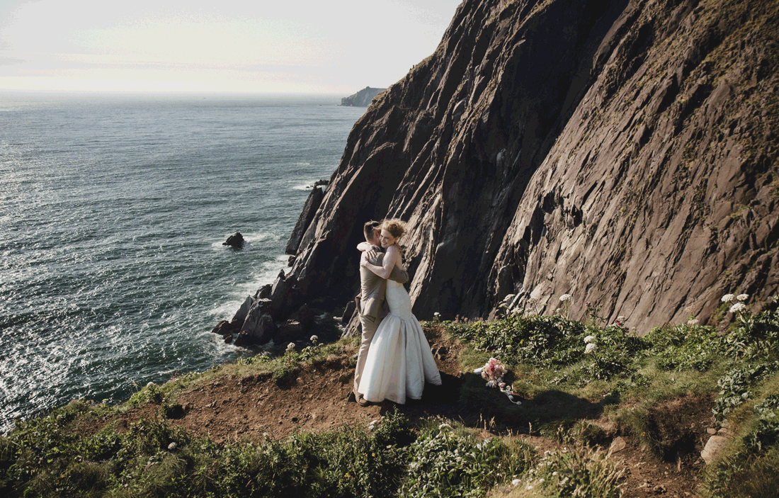 Couple in wedding attire embracing on dramatic coastal cliffs with ocean waves and rocky shoreline backdrop.