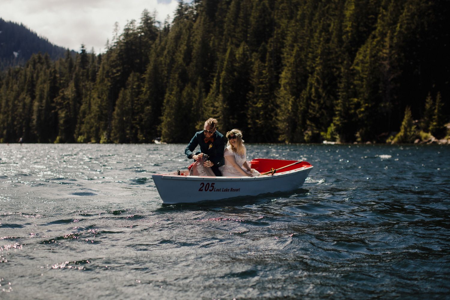 Couple rowing small white boat on mountain lake surrounded by pine forest.