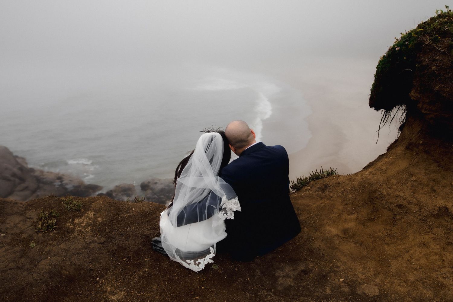 Bride and groom sit together on a misty coastal cliff, wedding veil blowing in the wind, overlooking the foggy ocean.