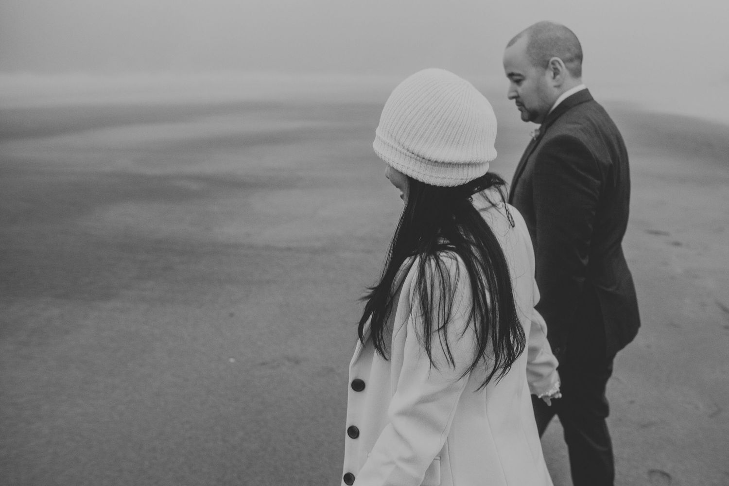 A couple holds hands on a misty beach, woman in white coat and knit hat, man in dark suit.