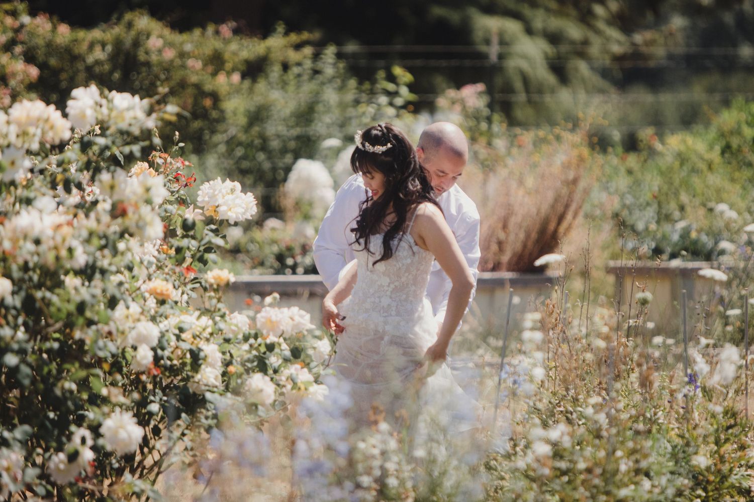 Bride and groom walking among blooming white roses in a lush garden during their outdoor wedding ceremony.