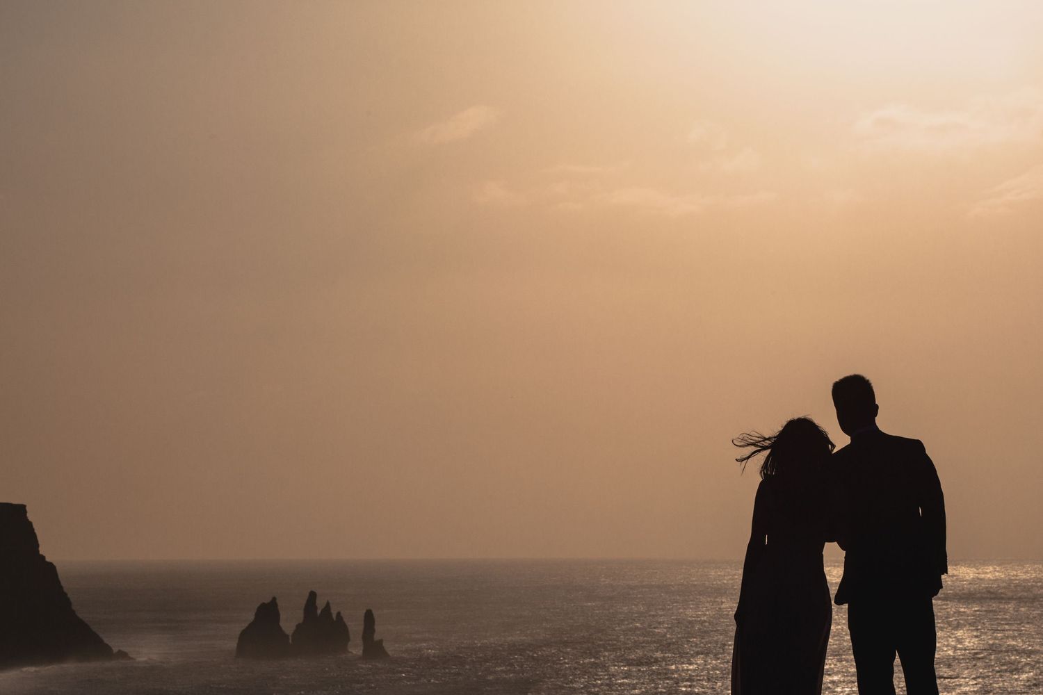 Two silhouettes stand close together against a golden sunset sky over the ocean.