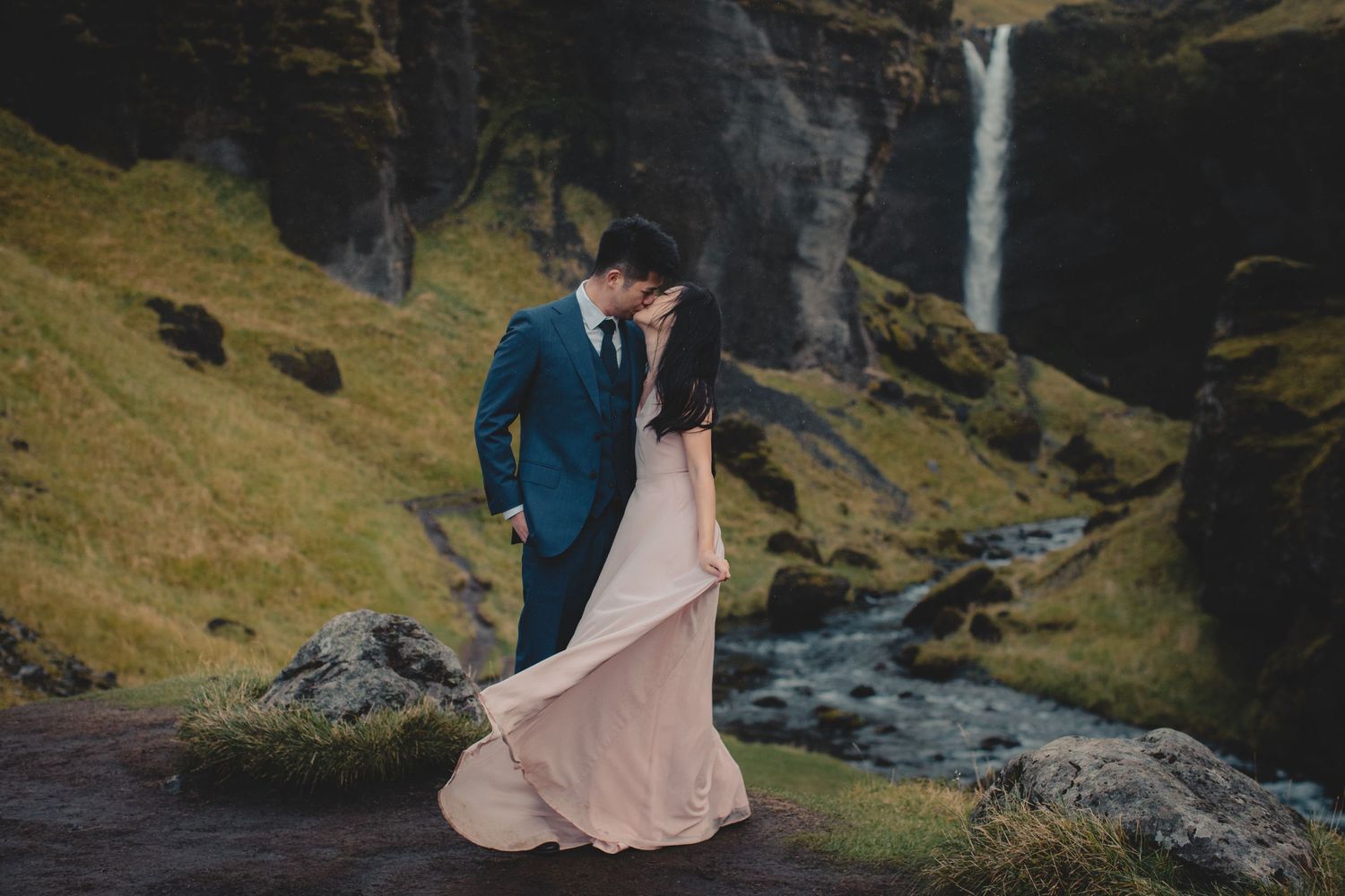 Couple in formal attire poses near dramatic waterfall and mountain stream.
