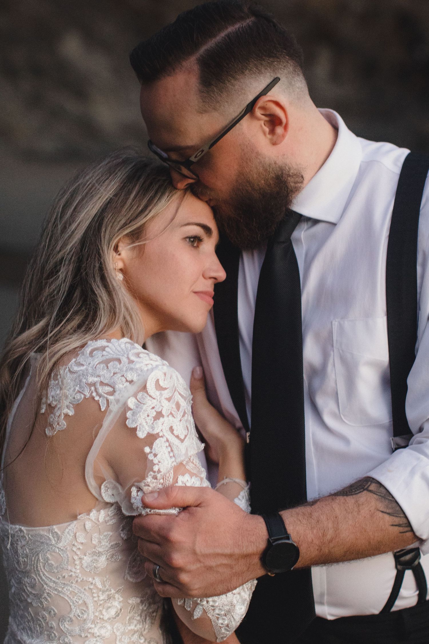 Bearded groom in suspenders tenderly embracing bride in lace wedding dress during intimate portrait session.