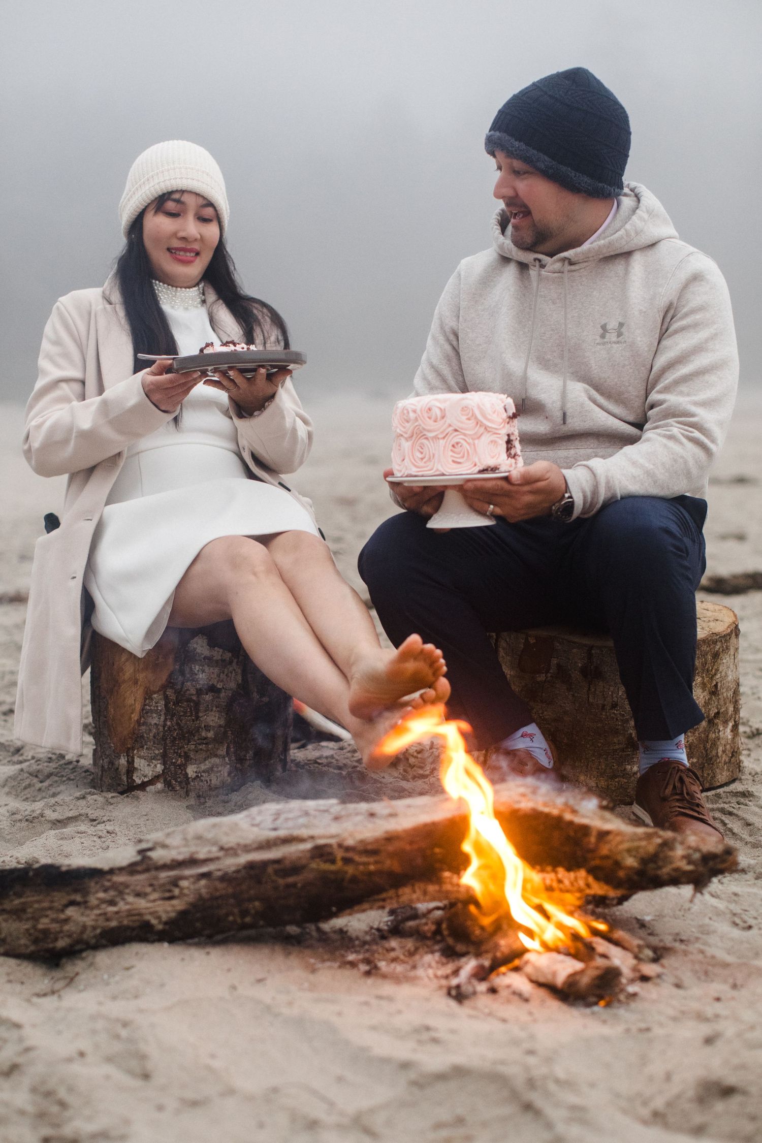 Couple sits by a beach bonfire holding pink wedding cake slices in foggy weather.