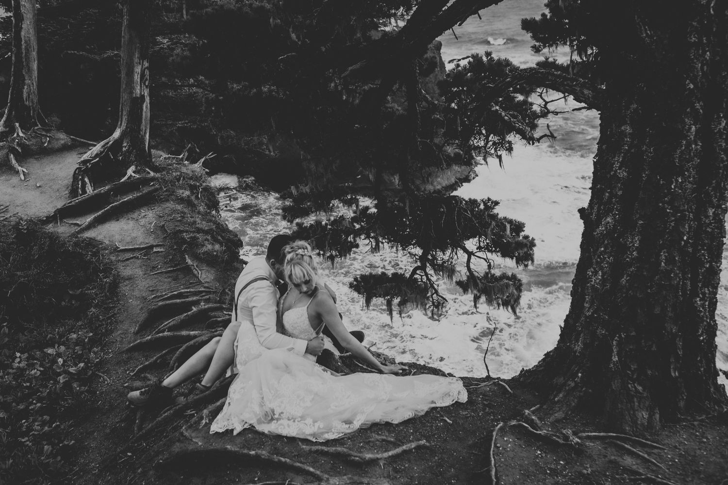 A black and white wedding portrait shows a couple resting among forest trees with ocean views.