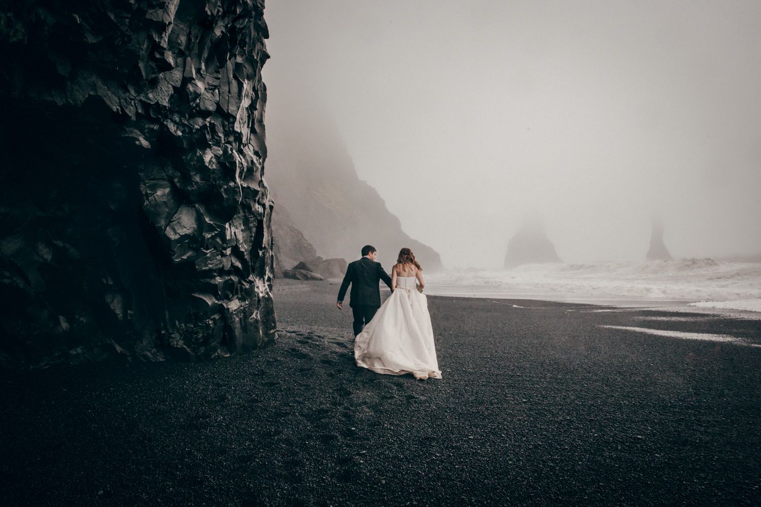 Couple walking along a misty black sand beach with towering cliffs in Iceland.