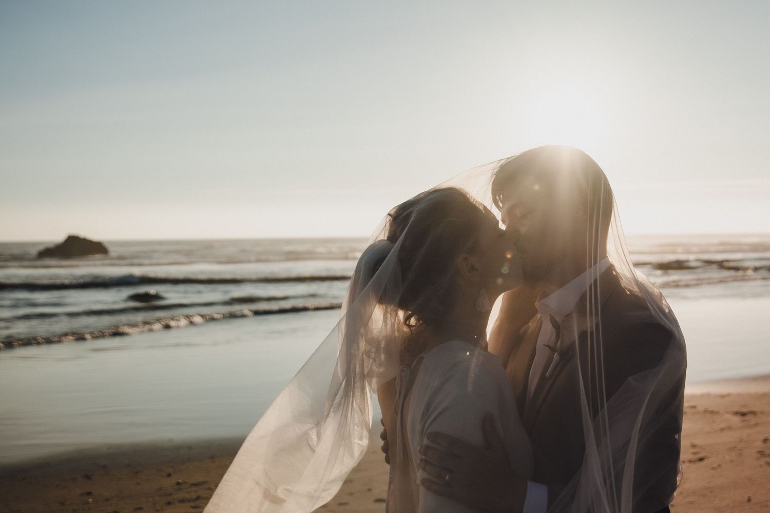Couple embracing under a sheer veil on a sunlit beach at sunset, with ocean waves and a rock formation in the background.