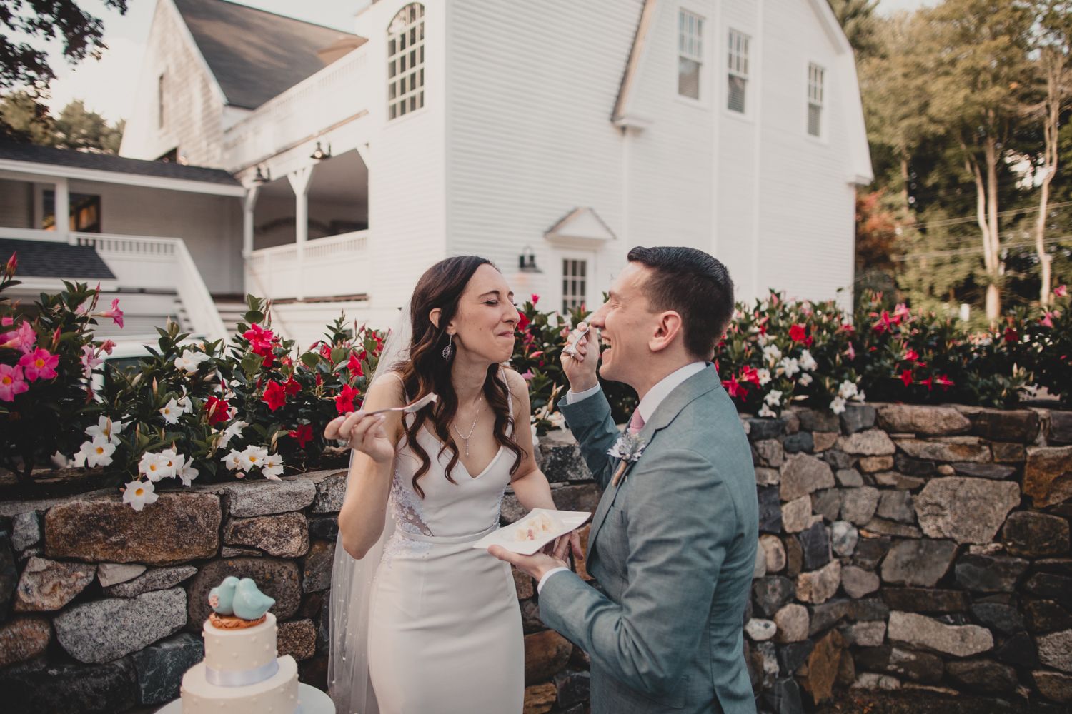A joyful couple shares a playful moment while tasting wedding cake in front of a white house with stone wall and roses.