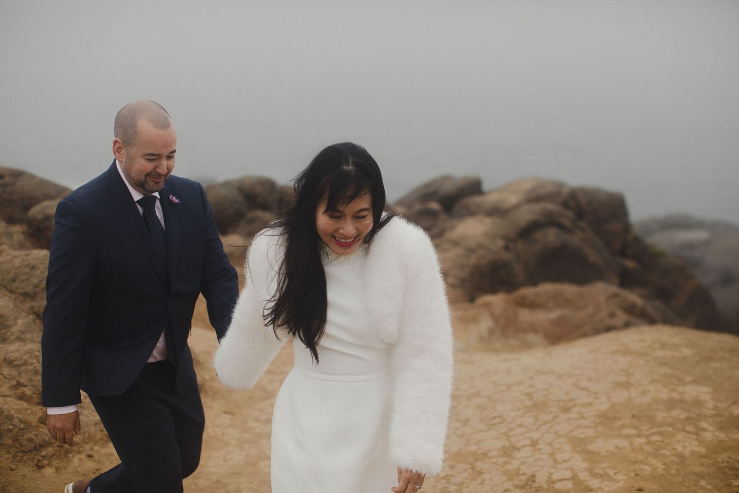 Couple walking hand in hand along a misty coastal rocky shoreline.