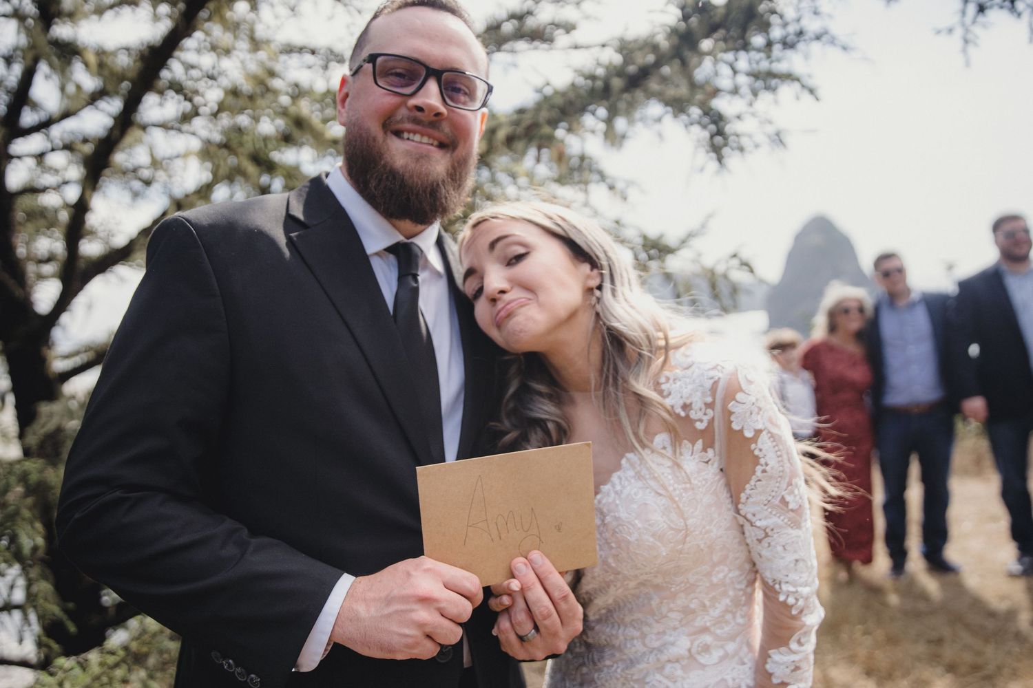 Smiling bride in lace gown leans on groom in black suit holding a card at outdoor ceremony.
