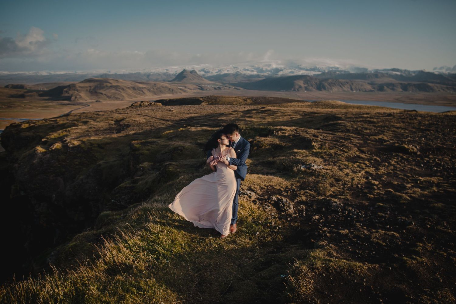 A couple poses together on a windswept highland overlooking snow-capped mountains.