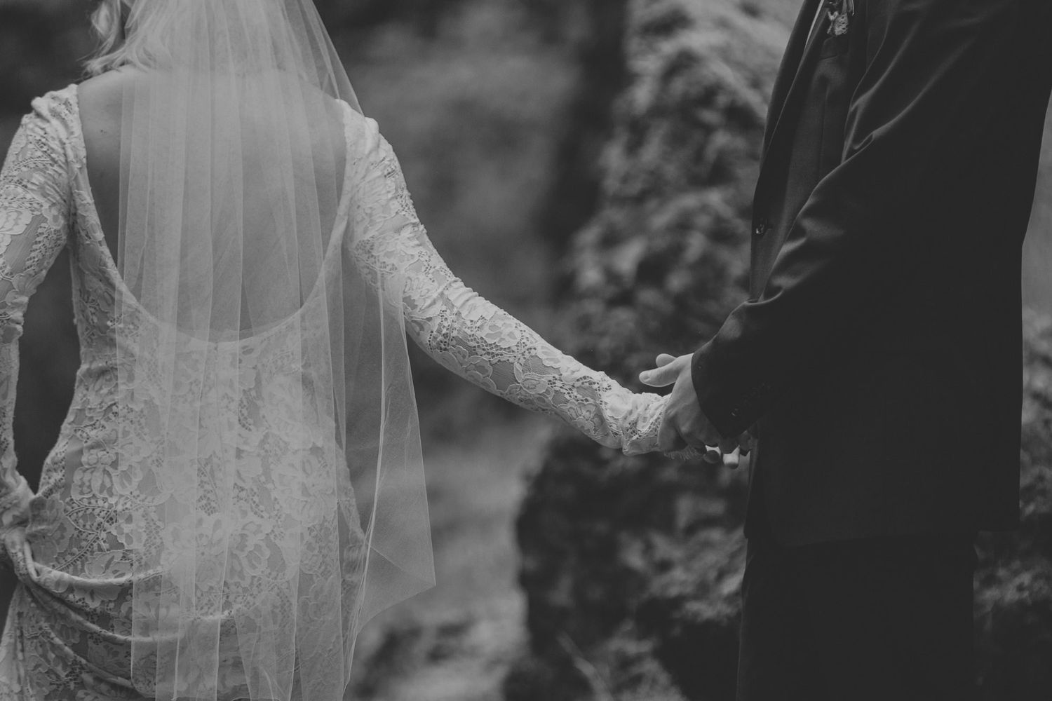 Black and white image of a bride in lace dress holding hands with groom against blurred background.