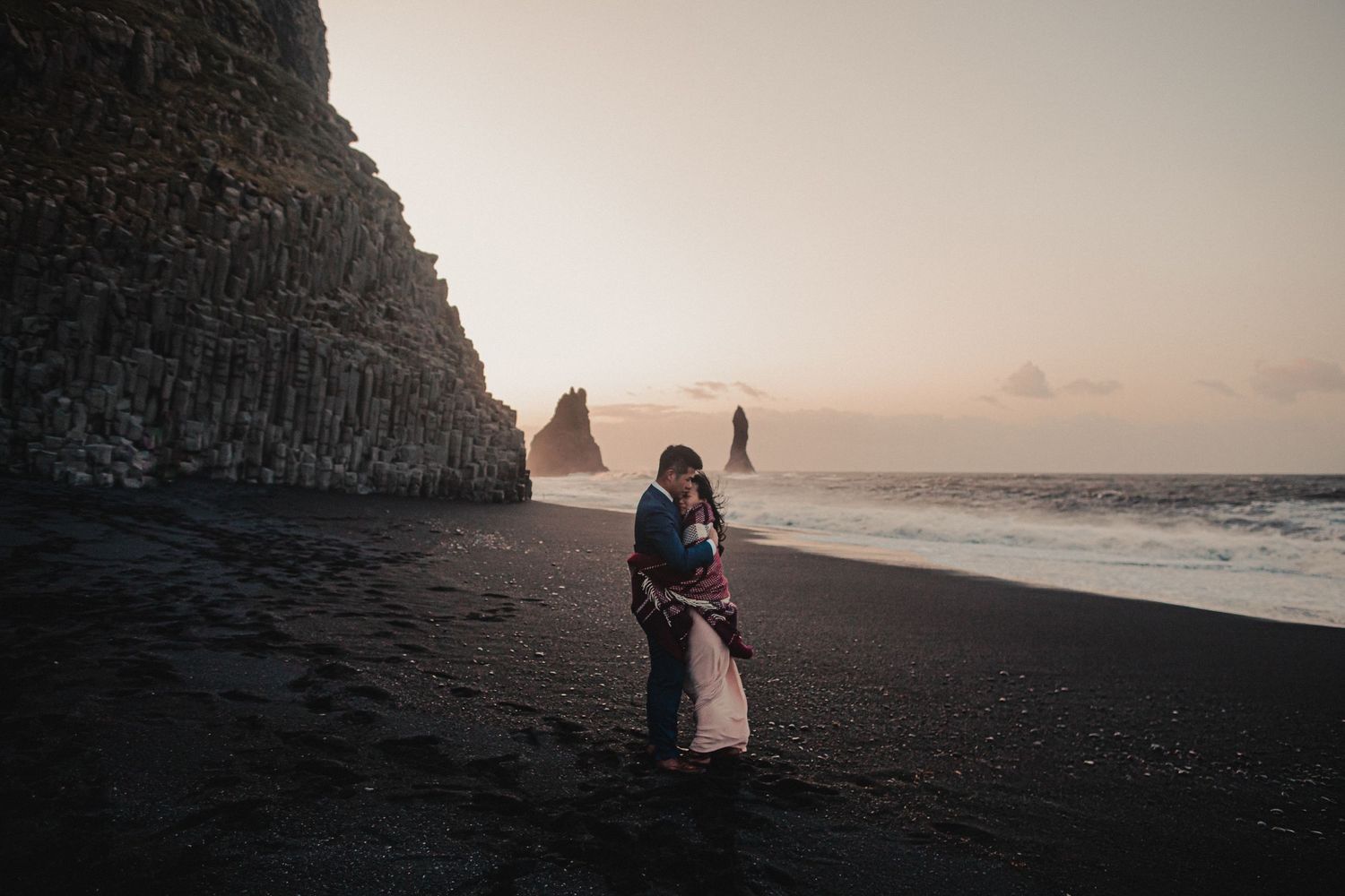 A couple embraces on a black sand beach with towering cliffs and sea stacks at sunset.