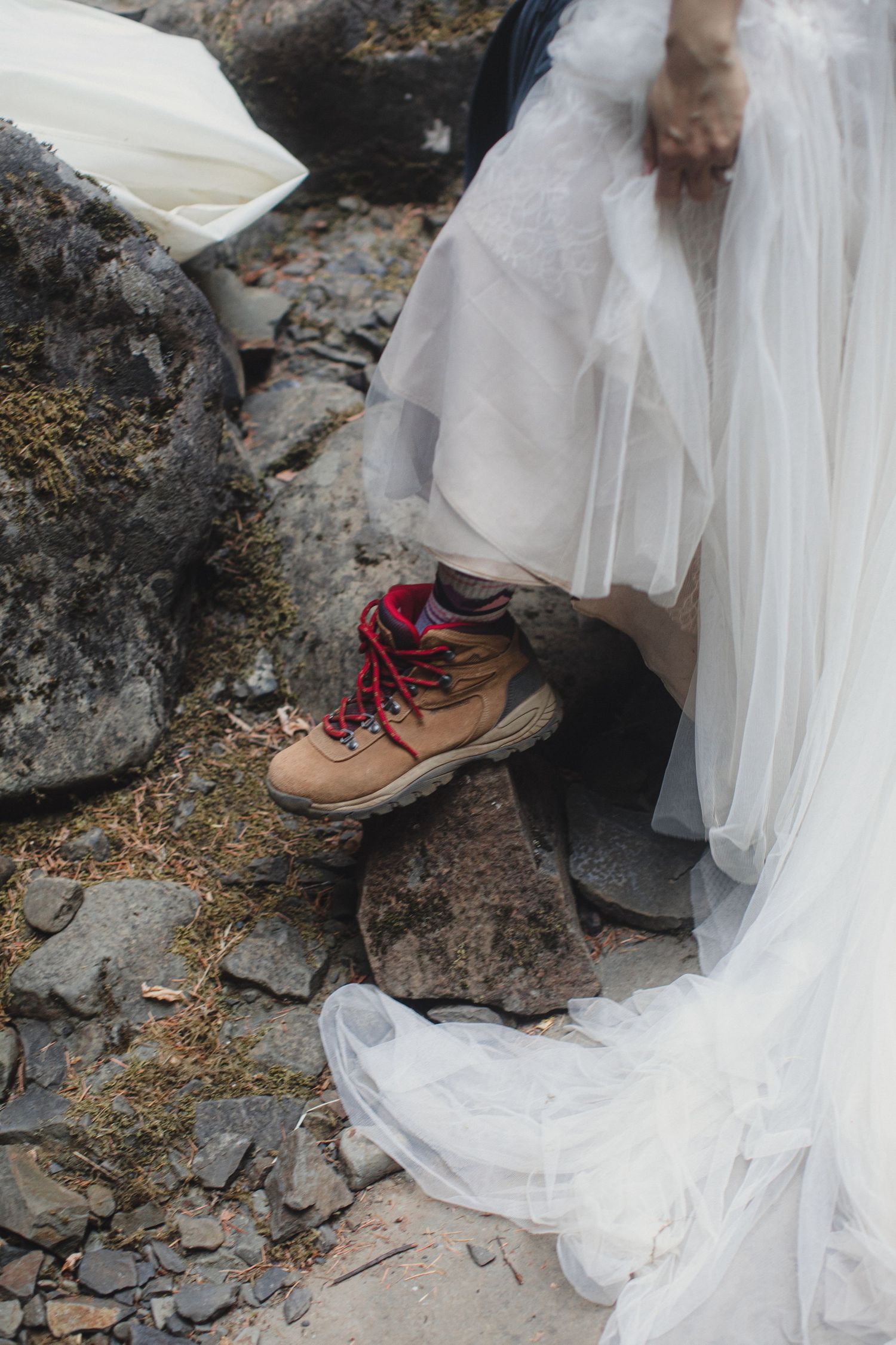 Bride wearing hiking boots under white tulle wedding dress on rocky terrain.