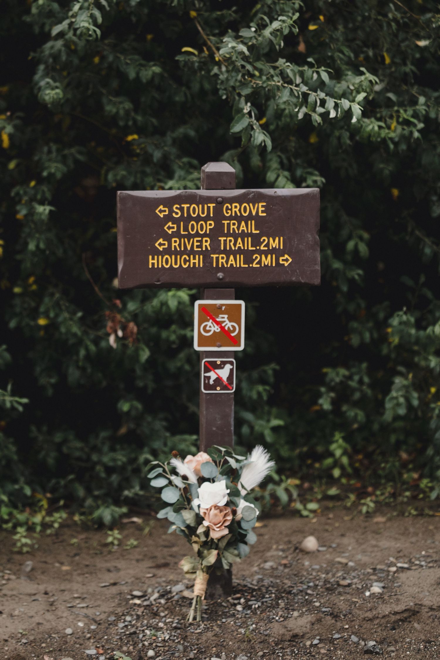 A wooden trail sign for Stout Grove Loop and Hiouchi Trail adorned with a bridal bouquet of pink and white flowers.