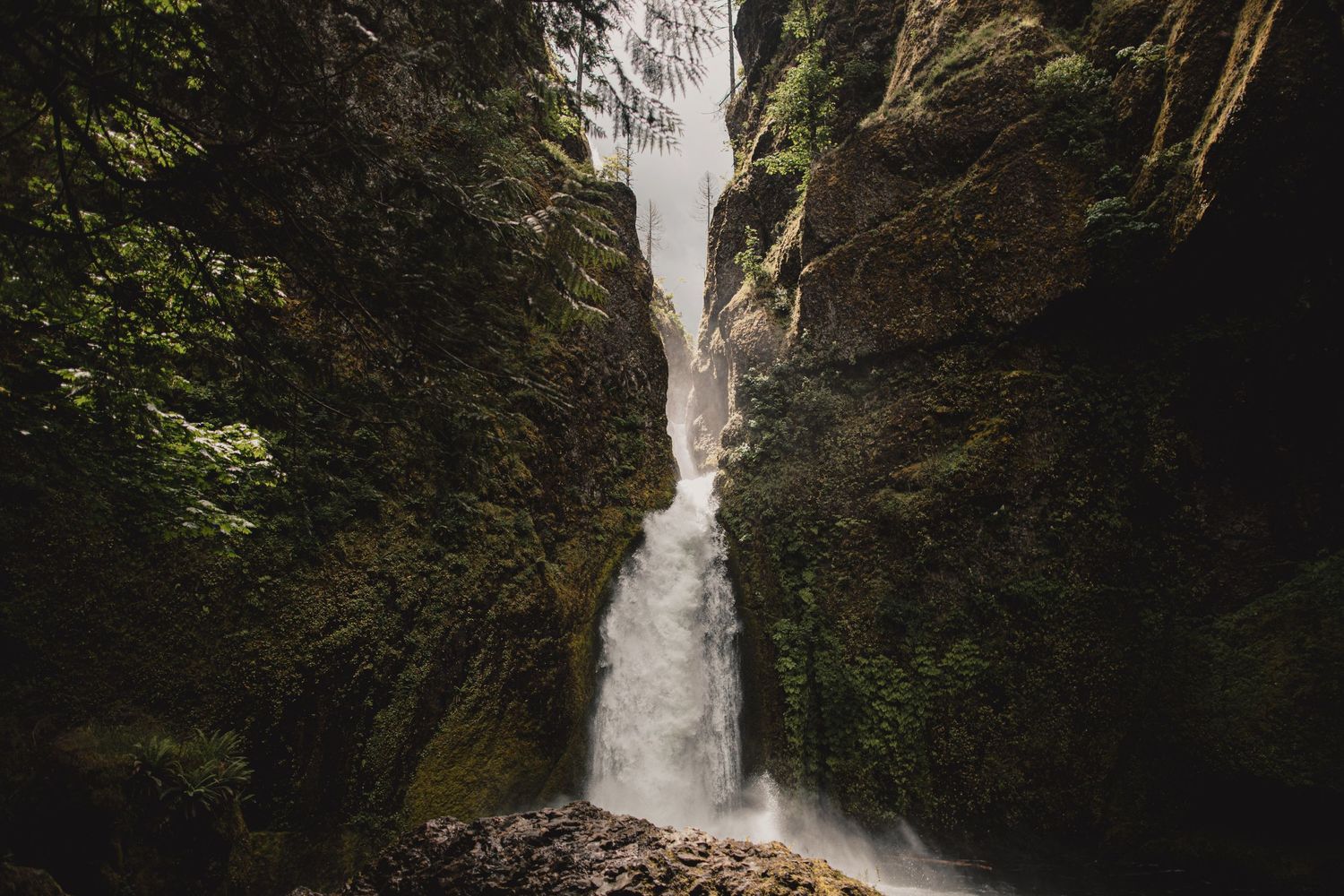 A dramatic waterfall cascades down moss-covered rock walls in a narrow canyon.