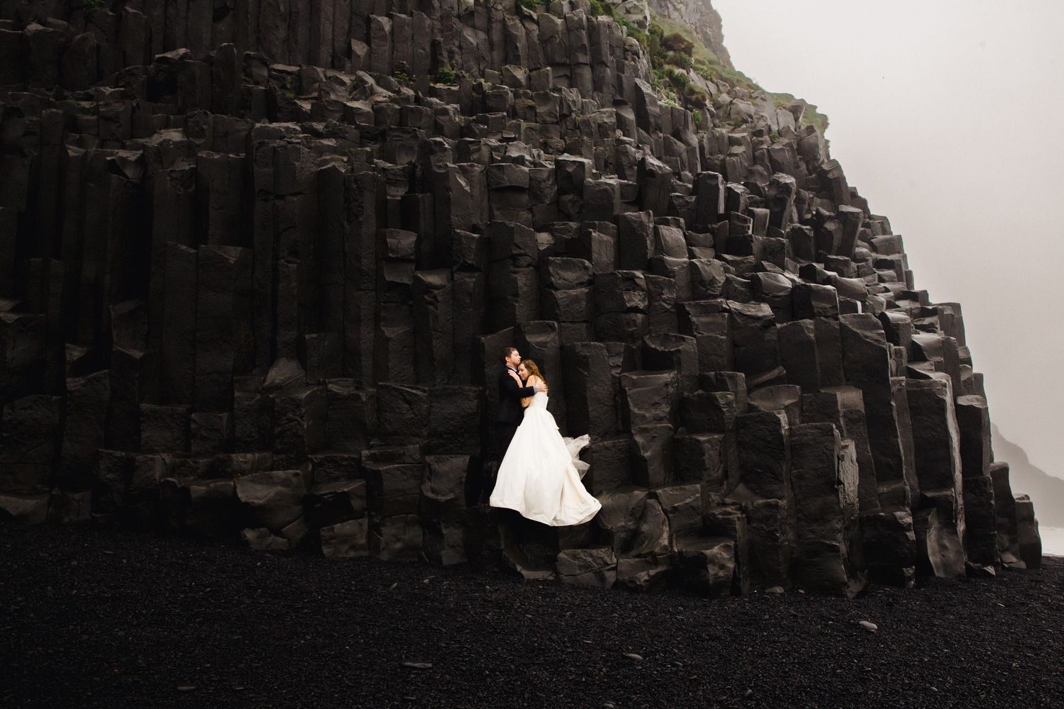 Bride in white gown poses against dramatic black basalt columns on Iceland's volcanic beach.