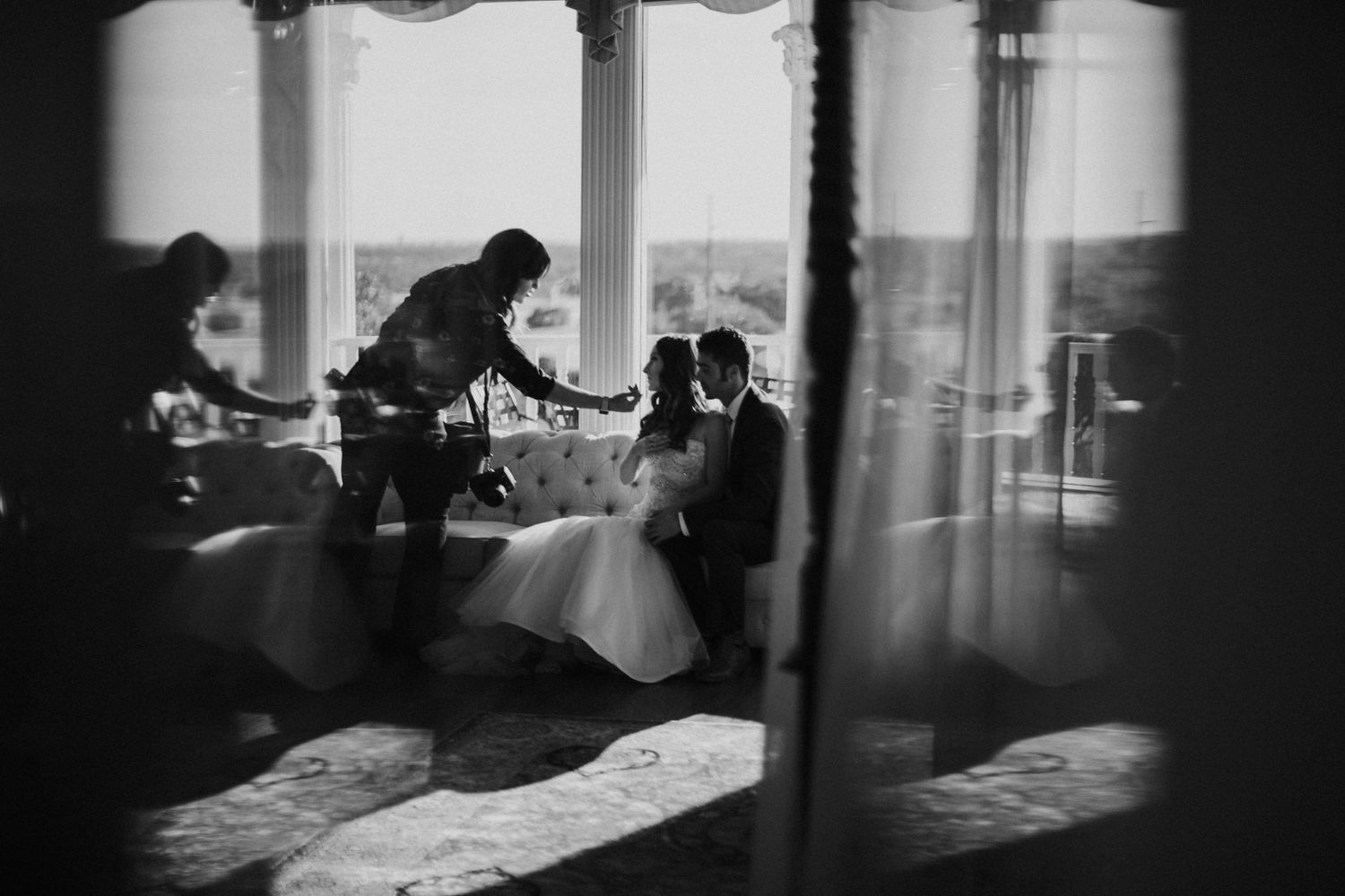 Black and white wedding photo of bride and groom seated while a photographer captures their moment in an elegant venue.