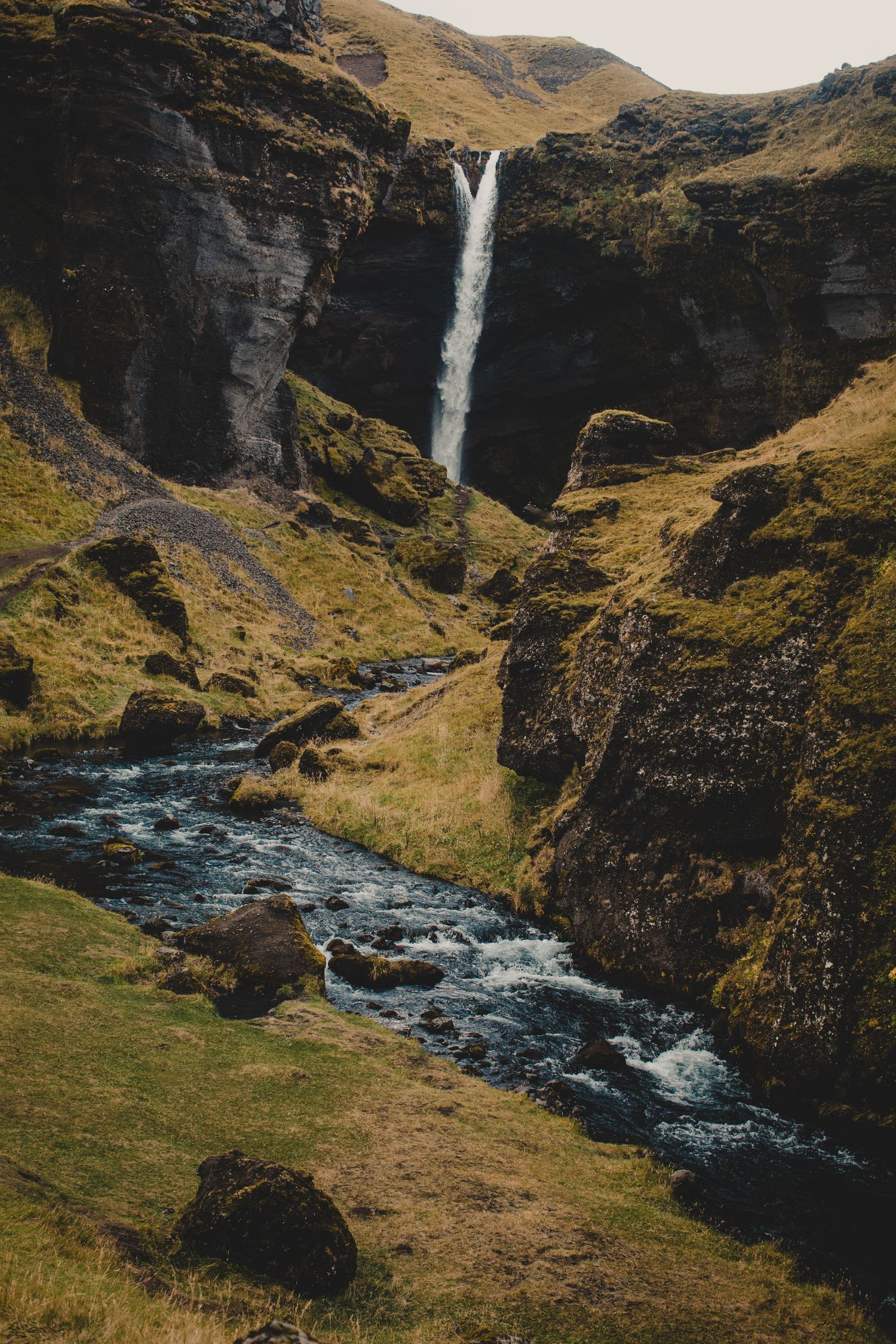 Dramatic waterfall cascades down dark cliffs with rushing river and green slopes in Iceland.